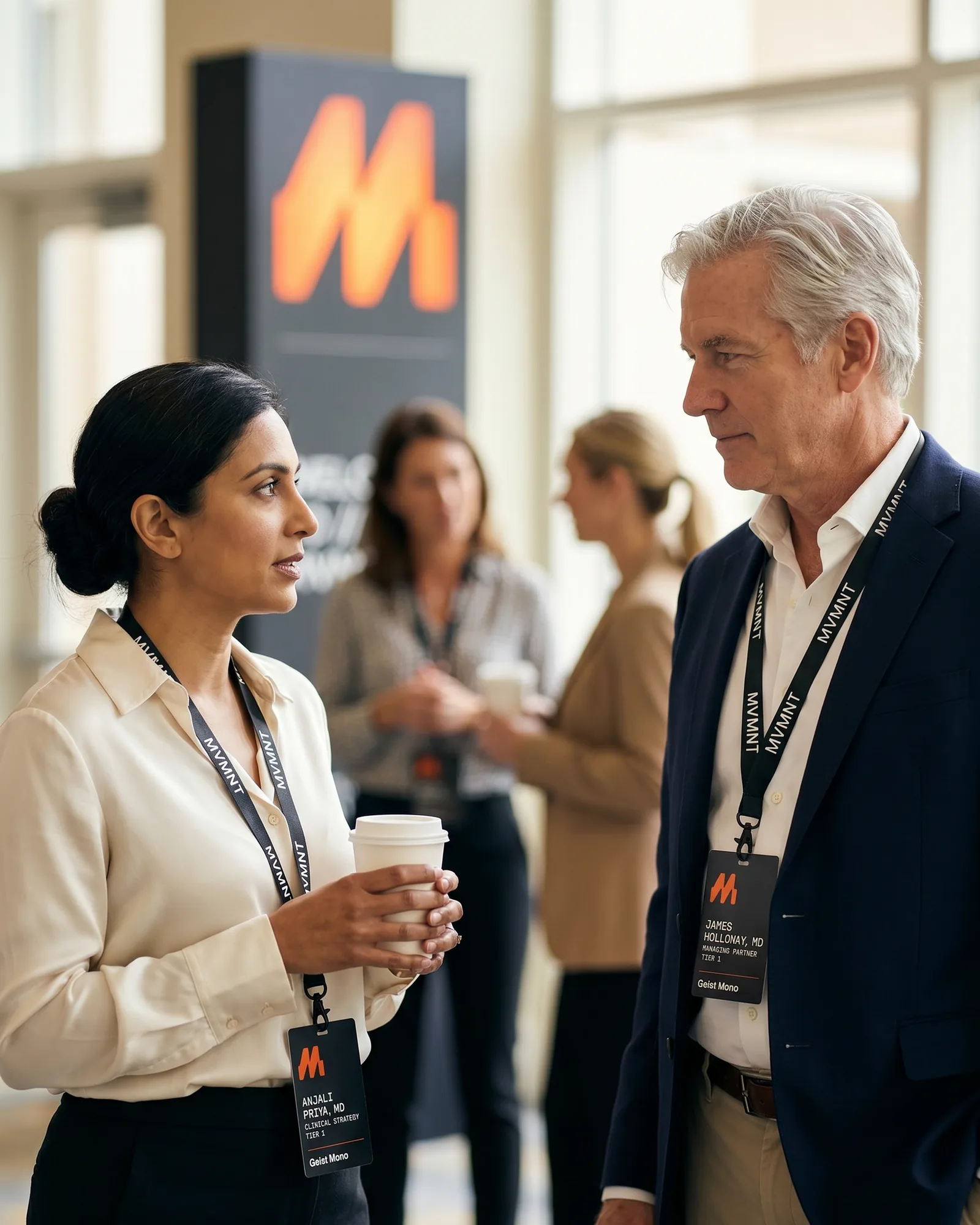 Two senior physicians in a summit lobby between sessions, mid-conversation as one finishes asking a clinical question.