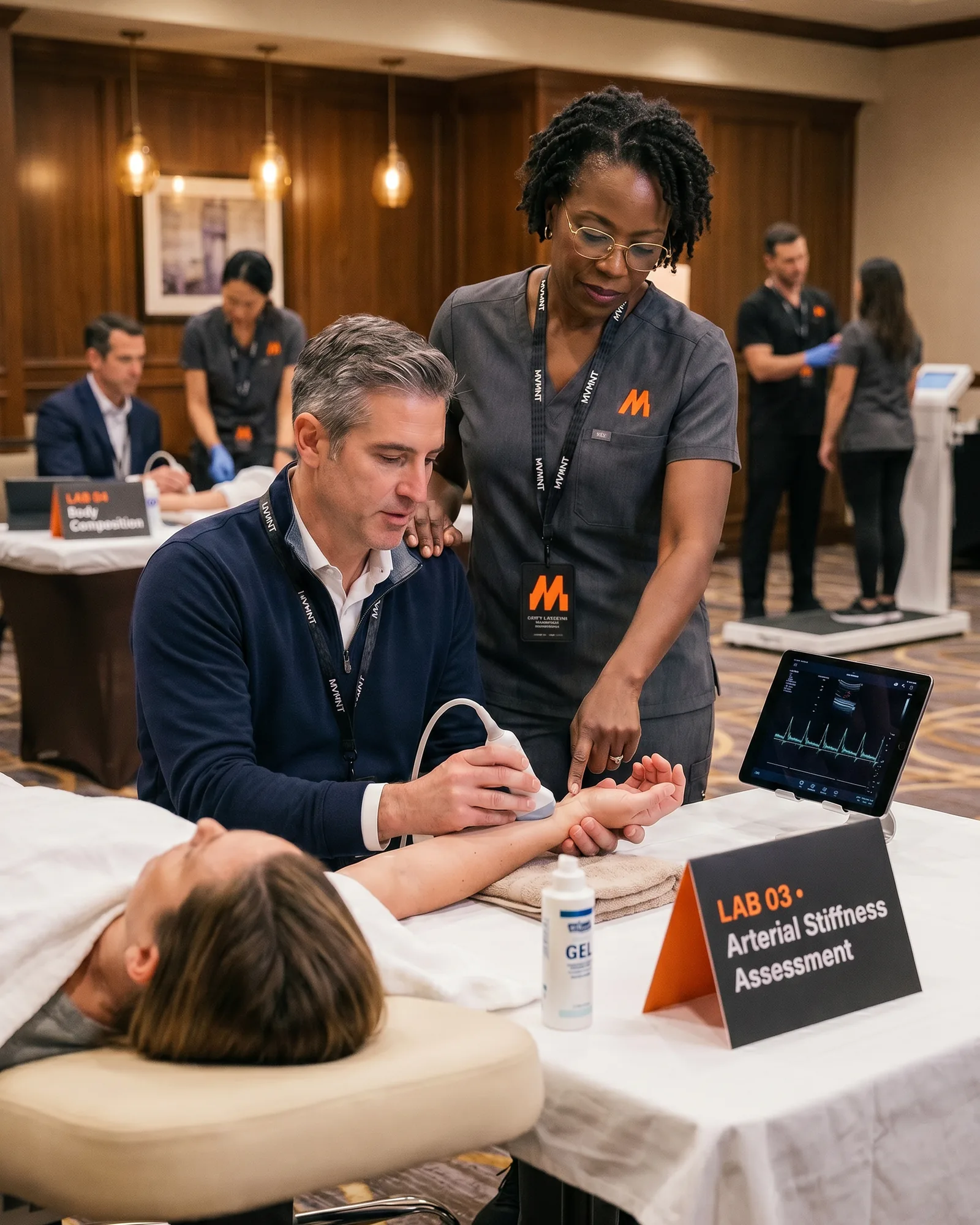 An instructor in MVMNT scrubs supervises a physician performing a point-of-care vascular ultrasound on the MVMNT lab floor.