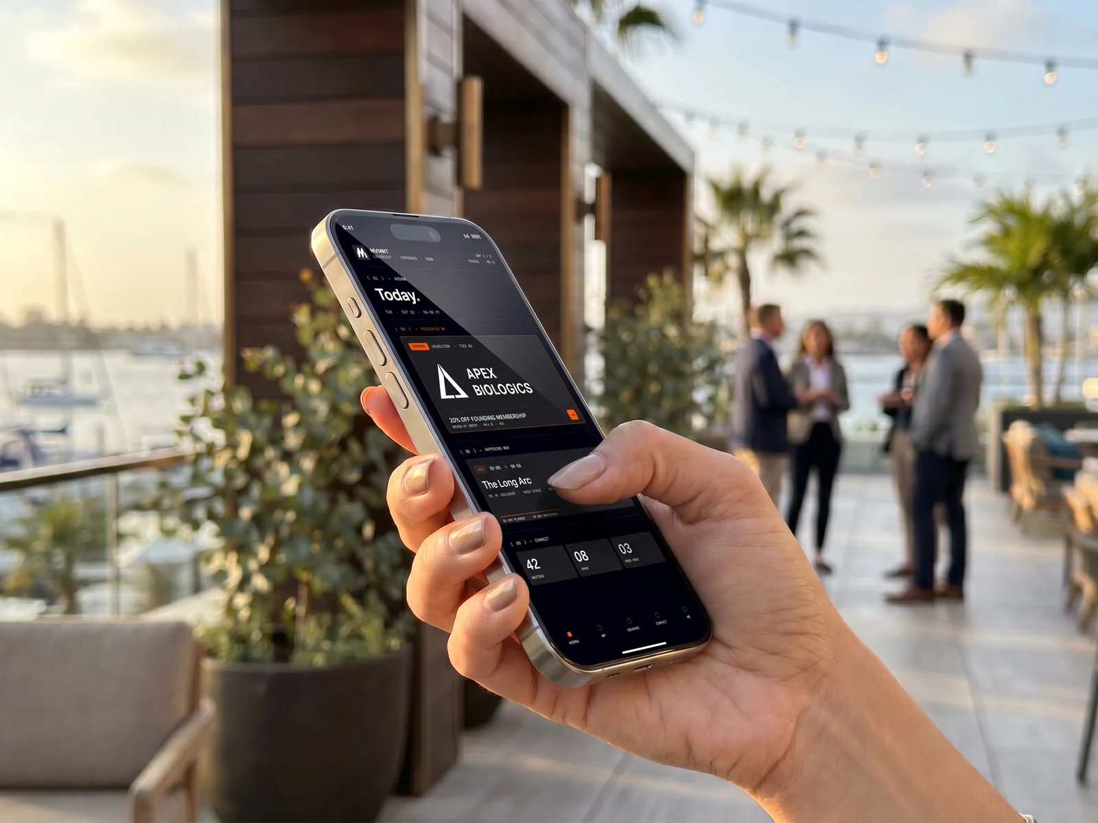 A hand holding a phone displaying the MVMNT meeting app, an Apex Biologics banner ad above the day's schedule; three attendees converse on a terrace overlooking the Coronado marina at golden hour.