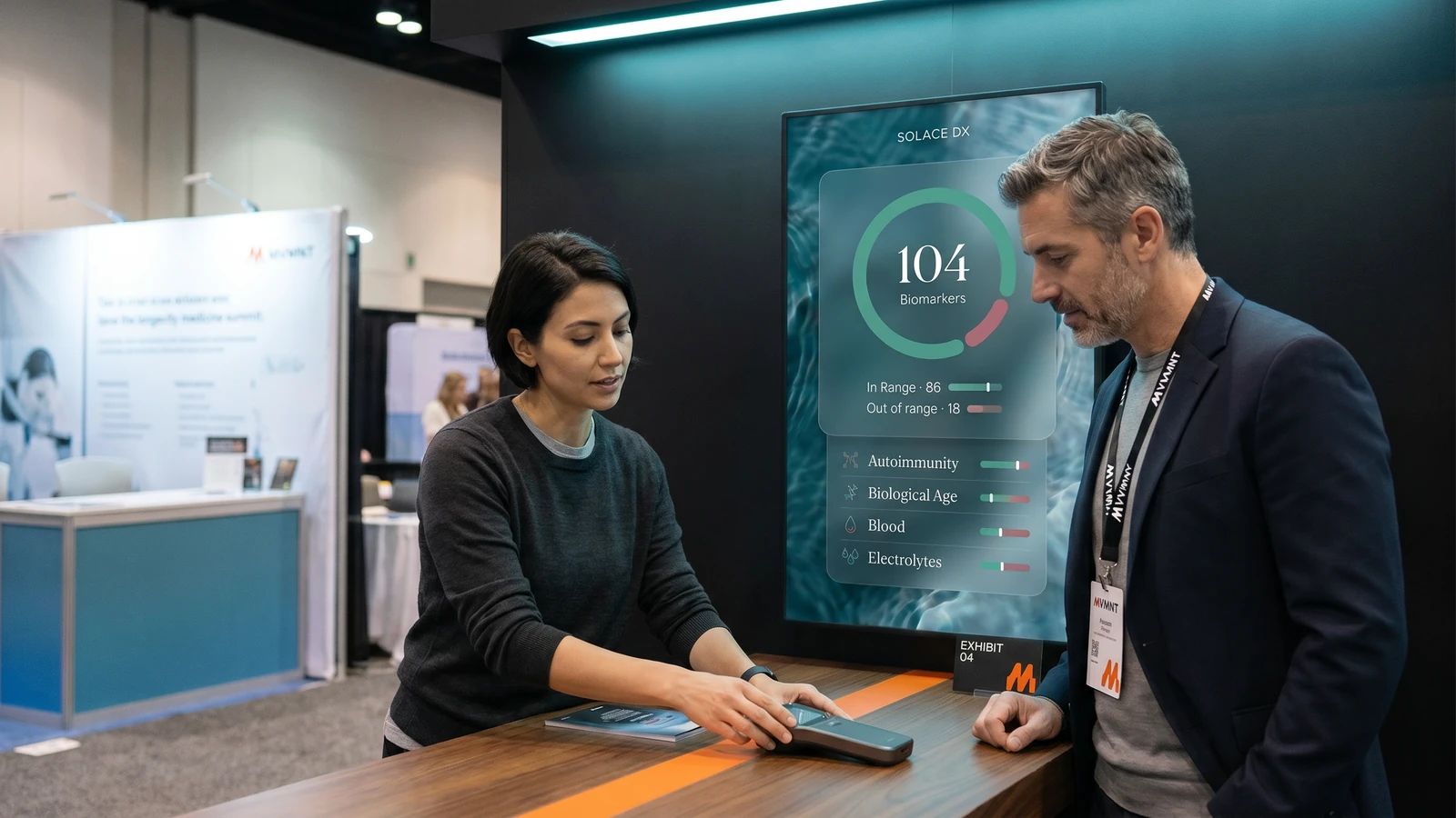 Two attendees engaged with a diagnostic device at an MVMNT exhibitor booth, product UI screen above.